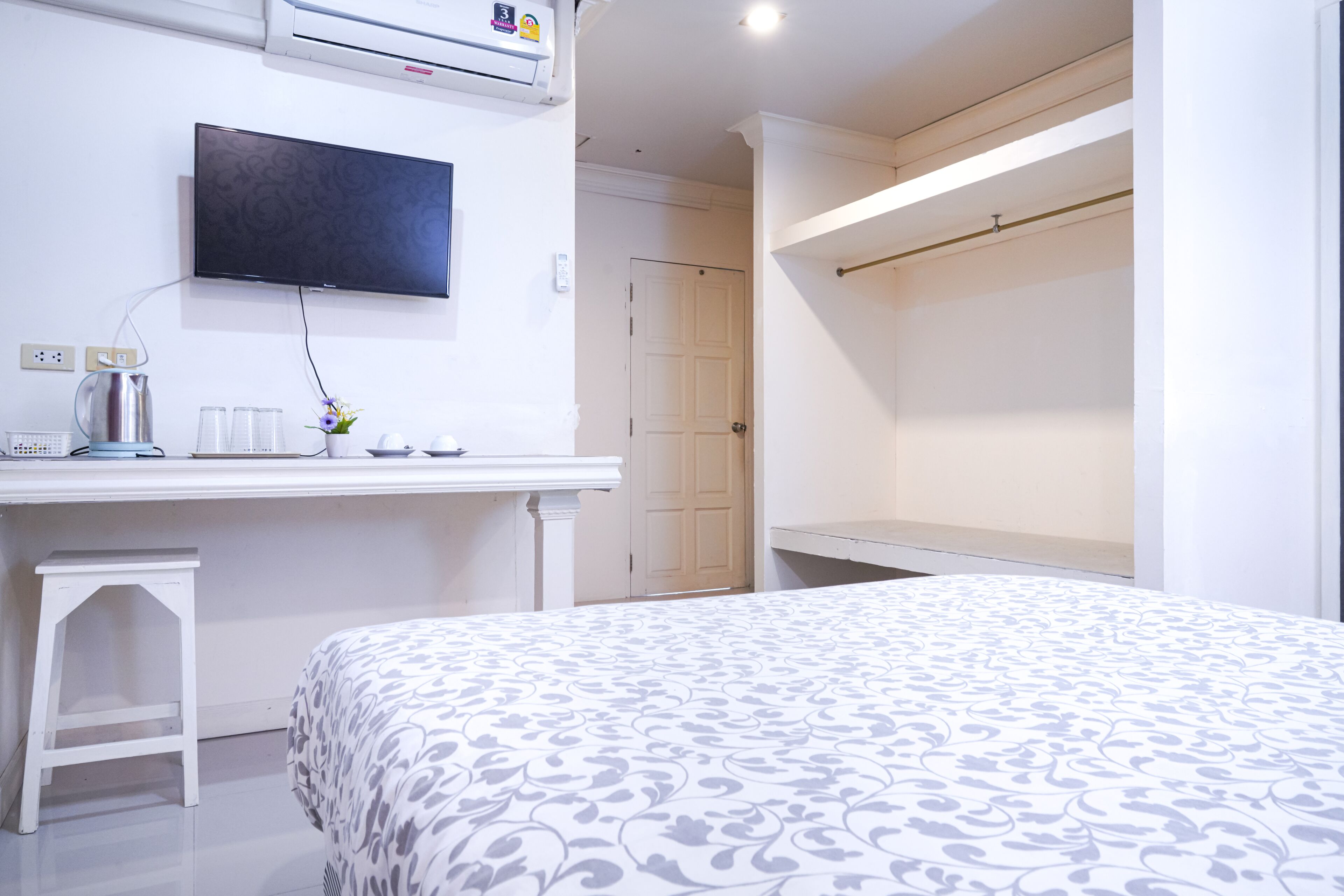 A bedroom with a bed, a TV mounted on the wall, a small table with a kettle and cups, and a door leading to another room.