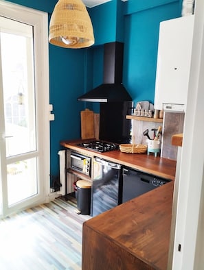 A compact kitchen with a gas stove, oven, and dishwasher, a black range hood, and a wicker pendant light.