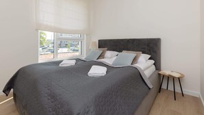 A modern bedroom with a large bed, a bedside table, and a window with a view of the outside. - Family Apartment with Parking | Balcony and Workspace (Warszawa)
