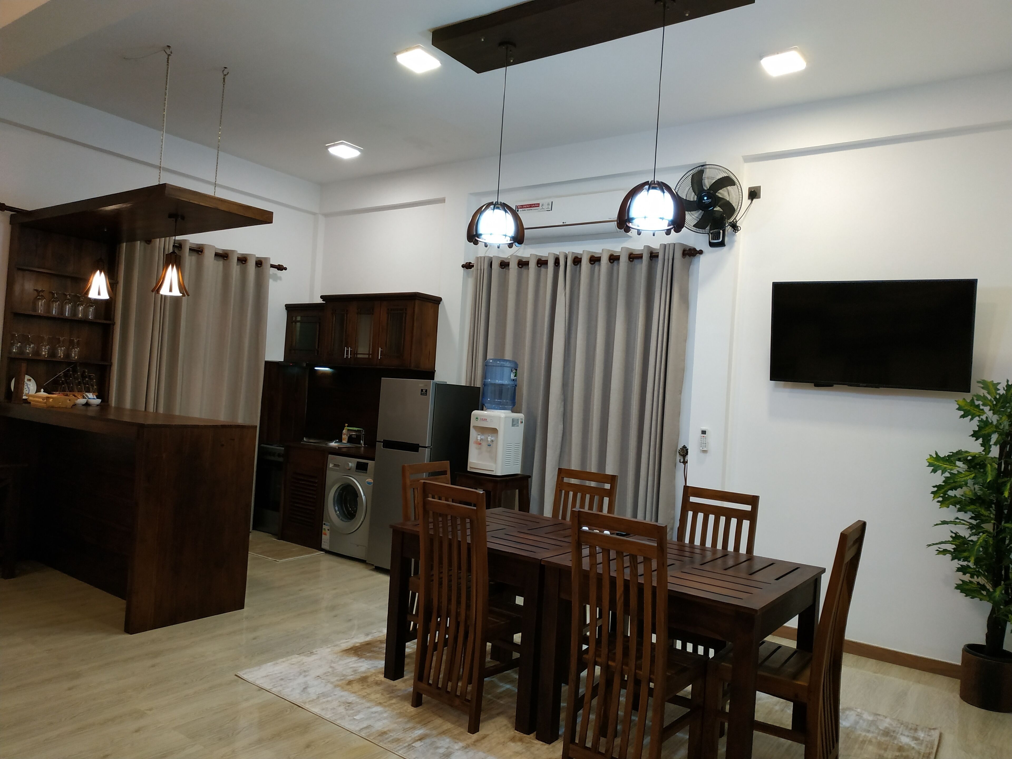 A modern kitchen with a built-in washing machine, a dining area with wooden furniture, and a flat-screen TV mounted on the wall.