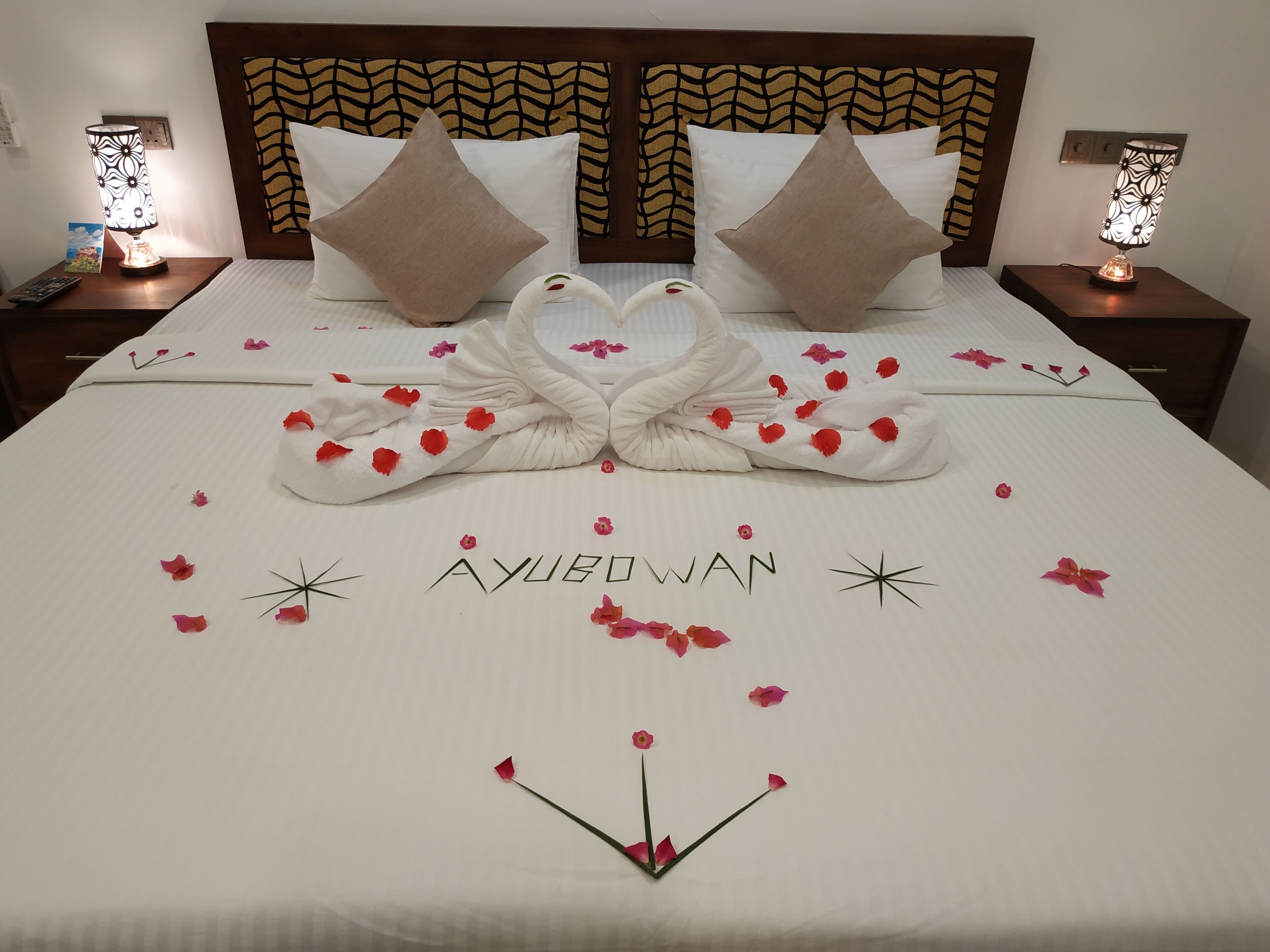Una cama impecablemente hecha con una composición de toallas en forma de cisne, rodeada de pétalos de rosa, y el nombre 'AYUBOWAN' escrito en la colcha.