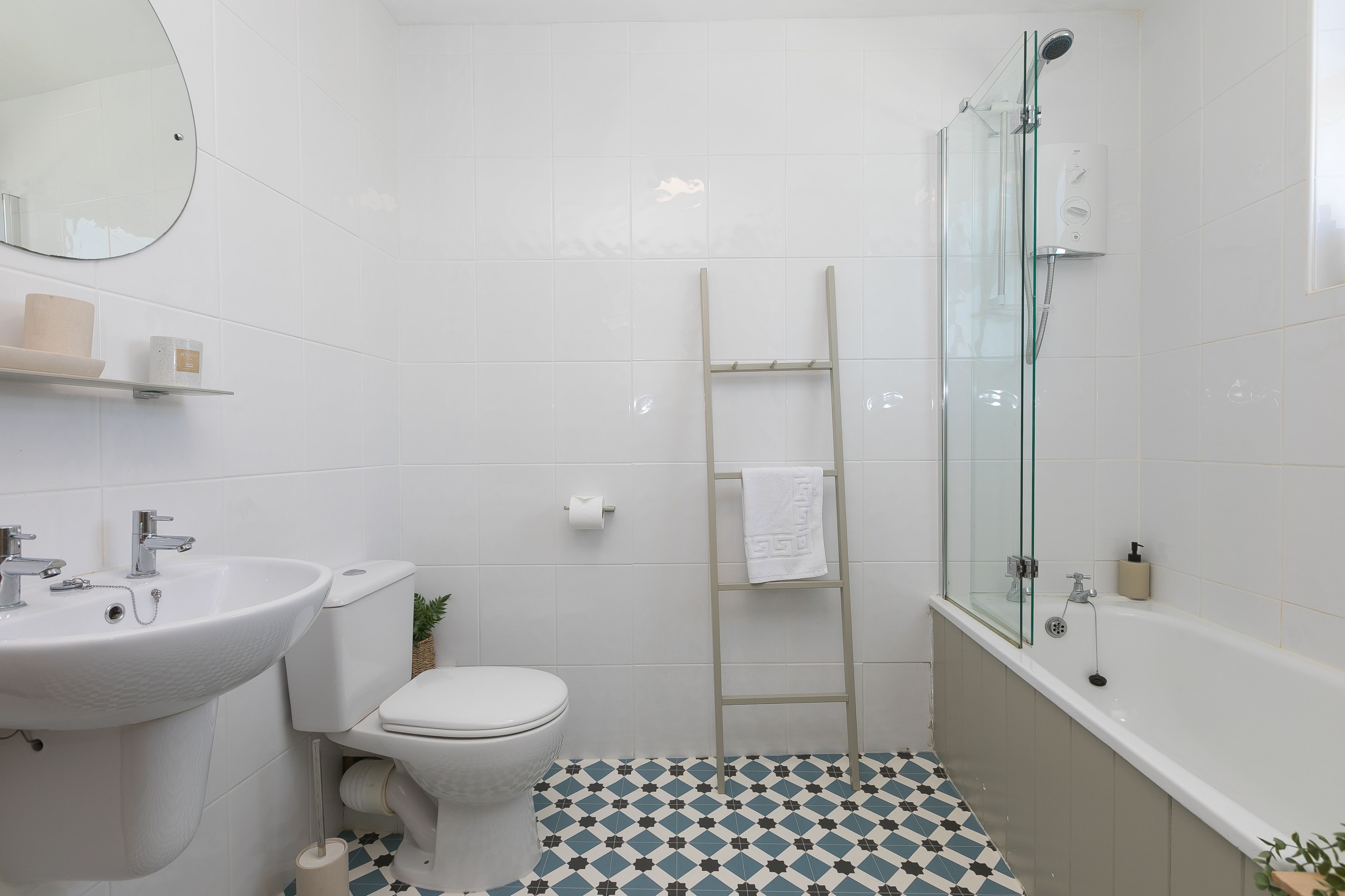 Et badeværelse med hvid vask, toilet og brusekabine med glasvægge. Der er monteret et håndklædetørrestativ på væggen, og gulvet har et mønster.