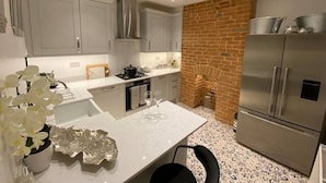 A modern kitchen with grey cabinets, a white countertop, a stainless steel refrigerator, and a brick accent wall. - Gloucester Place Townhouse + Hot Tub (Cheltenham)