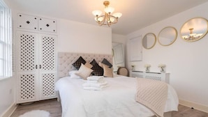 A bedroom with a tufted headboard, a large bed with white linens, a fluffy white rug, and a chandelier. - Gloucester Place Townhouse + Hot Tub (Cheltenham)