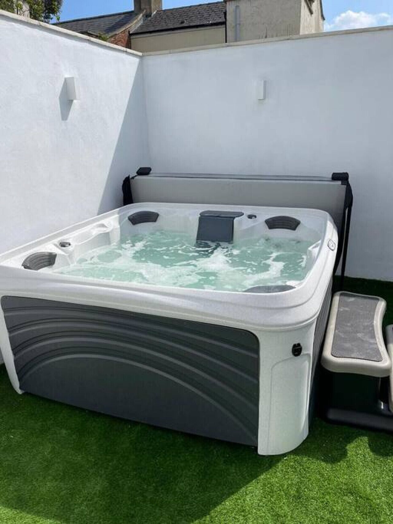 Un bain à remous doté d’un marchepied pour faciliter l’accès, entouré d’un mur blanc et d’une pelouse artificielle verte.