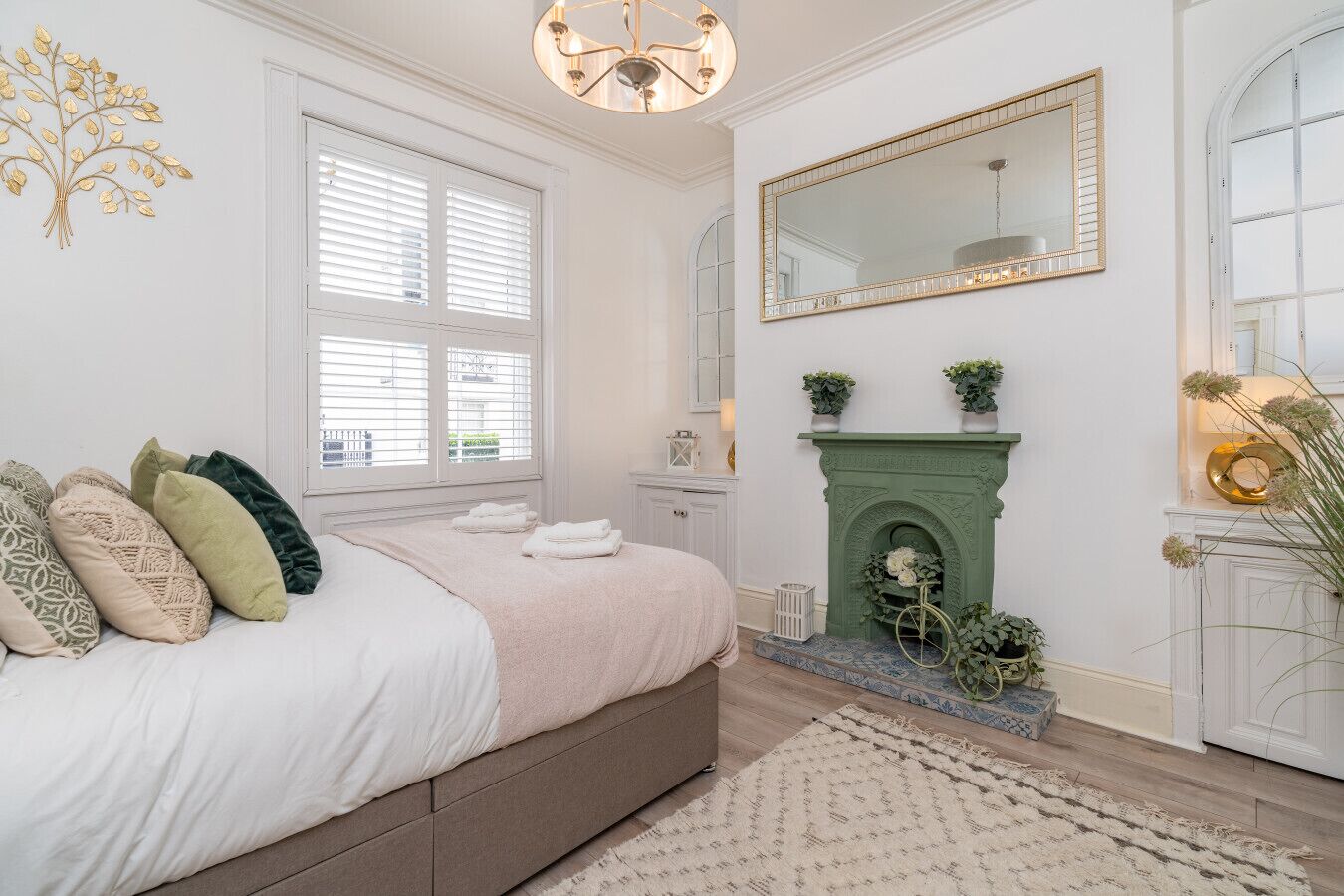 Une chambre à coucher avec un lit, une cheminée verte, un grand miroir et une fenêtre avec des stores.