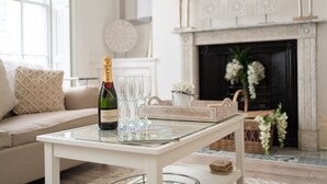 A living room with a sofa, a glass coffee table, a bottle of champagne, and glasses. There is a fireplace with decorative items and a mirror above it.
