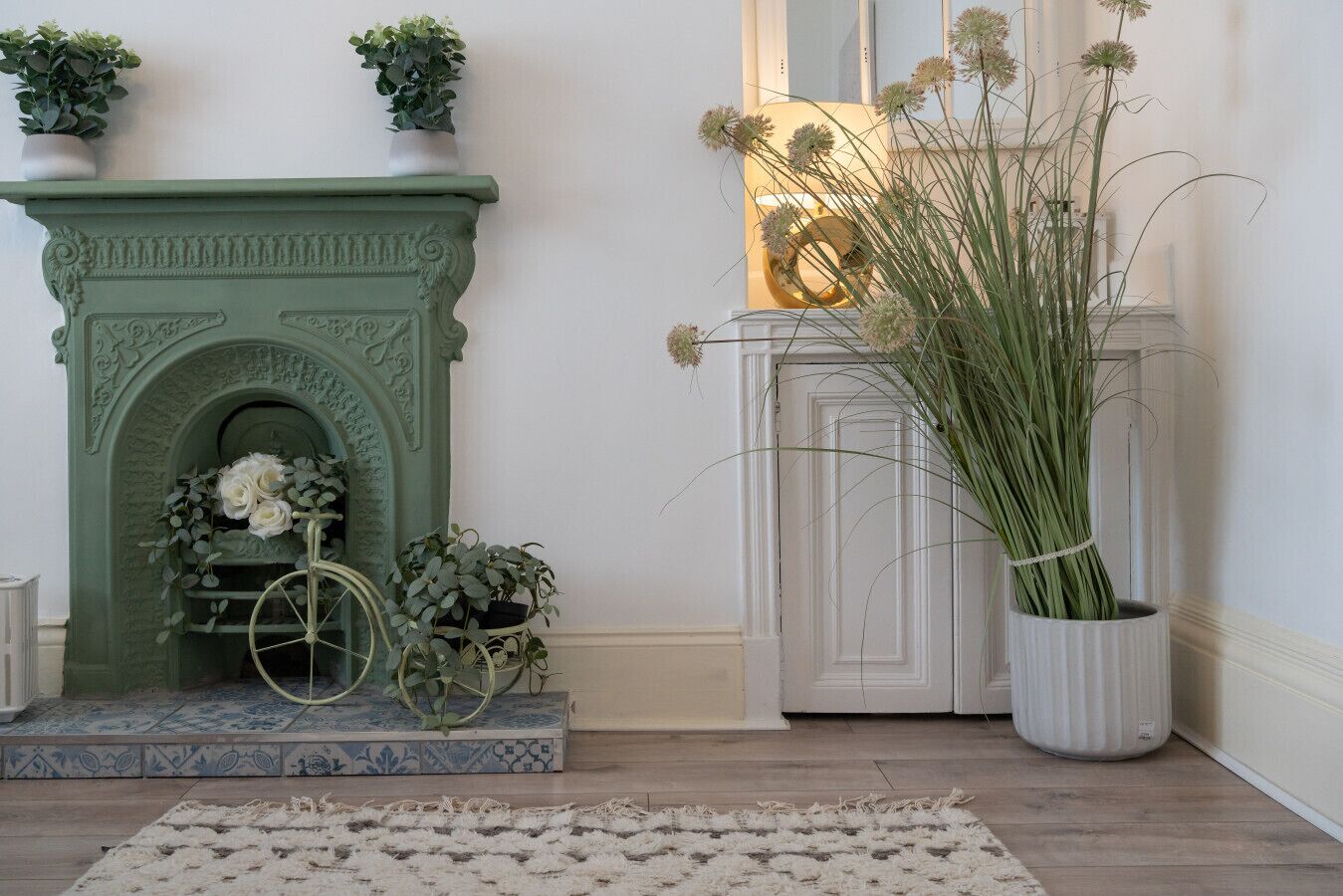 A green fireplace with a decorative wreath and a vintage bicycle, a white cabinet with a plant, and a large vase with tall green plants.