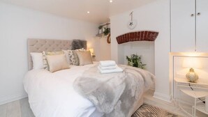 A bedroom with a tufted headboard, a bed with multiple pillows, a white wardrobe, a small table with a lamp, and a brick fireplace.