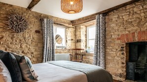 A rustic bedroom with a stone wall, a fireplace, a bed with pillows, and a hanging light fixture. - Windsor Cottage - Bolthole in the heart of CN (Chipping Norton)