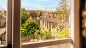 A view through a window of a stone building with a traditional roof, surrounded by greenery and a clear sky. - Windsor Cottage - Bolthole in the heart of CN (Chipping Norton)