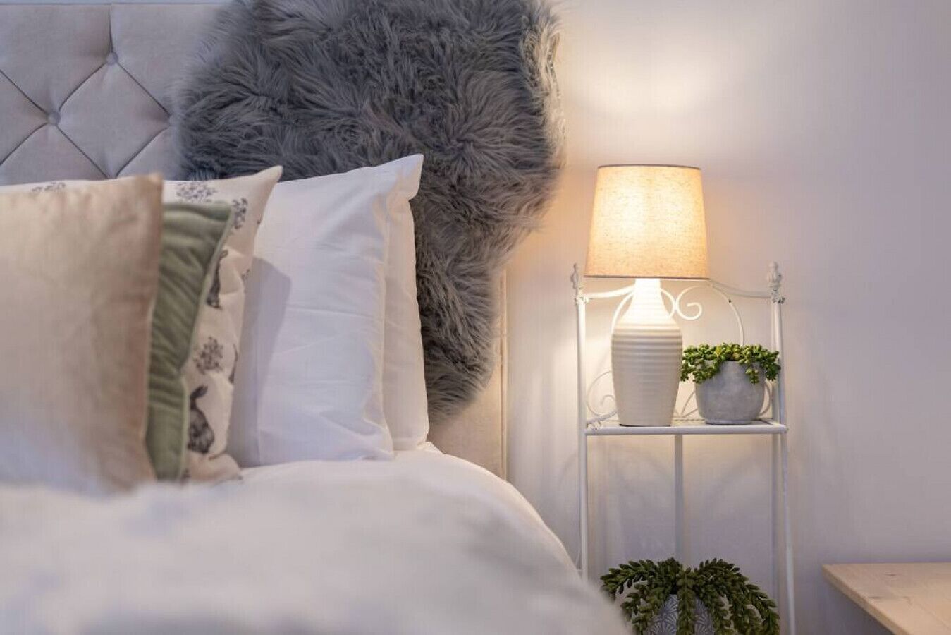 Un lit bien fait, agrémenté d’oreillers blancs et à motifs, d’une tête de lit moelleuse de couleur grise, et d’une table de chevet sur laquelle se trouvent une lampe et une petite plante en pot.