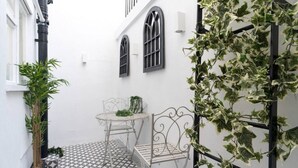 Une petite cour étroite avec un sol carrelé, une table et une chaise en métal, et un pot de fleurs.