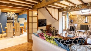 A cozy living room with a wooden ceiling, a sofa with colorful hexagonal cushions, and a dining area with wicker chairs. - Granary Cottage - Luxury Barn Conversion (Weston Subedge)