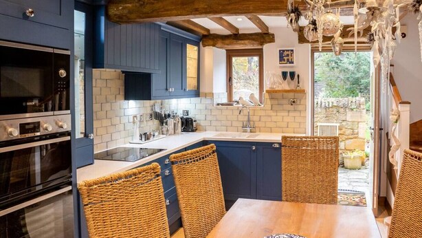 A modern kitchen with blue cabinets, a Bosch oven, and a dining area with wicker chairs. - Granary Cottage - Luxury Barn Conversion (Weston Subedge)