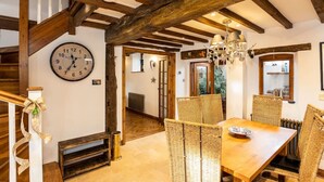 A dining area with a wooden table and wicker chairs, a clock on the wall, and exposed wooden beams. - Granary Cottage - Luxury Barn Conversion (Weston Subedge)