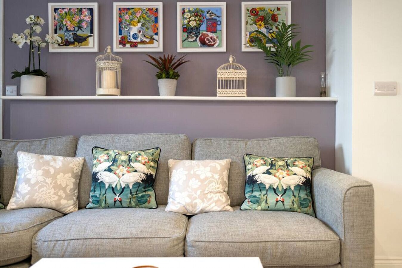 A grey sofa with floral-patterned cushions, a white coffee table, and a wall with framed floral paintings and decorative items.