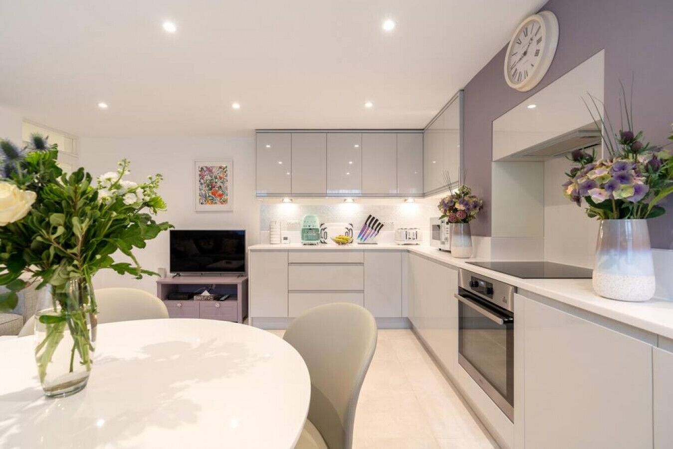 A modern kitchen with white cabinets, a built-in oven, and a dining area with a round table and chairs.