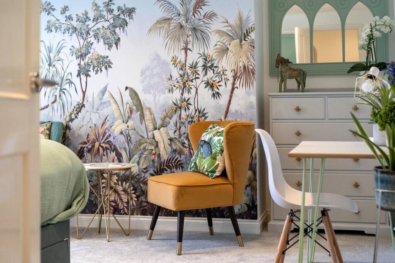 A room with a mural of tropical plants, a mustard-colored armchair with a floral pillow, a small round table, a white dresser with floral drawer knobs, and a mirror with a horse decoration.