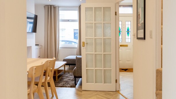A room with a dining table and chairs, a sofa, a door with frosted glass panels, and a view of the street through a window. - Bright & Airy 2BD - 10 mins walk into Centre (Cheltenham)