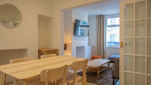 A modern living room with a wooden dining table, chairs, a TV mounted on the wall, a sofa, and a glass door leading outside. - Bright & Airy 2BD - 10 mins walk into Centre (Cheltenham)