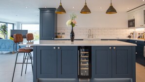 Een moderne keuken met donkerblauwe kastjes, een centraal kookeiland met wijnkoeler, hanglampen en uitzicht op de omgeving.