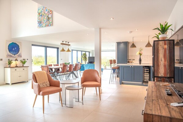 Een ruime woonkamer met een eettafel, twee leren fauteuils en een salontafel. Er is een keuken met blauwe kasten en een houten kookeiland.