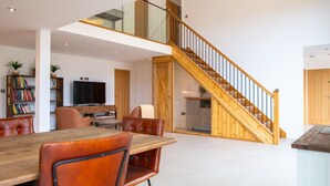 Een ruime woonkamer met een houten trap, een eettafel met stoelen, een televisie en een boekenkast.