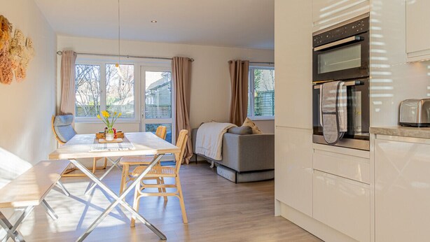 A modern kitchen with white cabinets, a built-in oven, and a toaster. There is a dining area with a table and chairs, and a sofa in the background. - Modern 2 BD w Garden in the Heart of Tetbury (Tetbury)