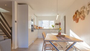 A modern kitchen with white cabinets, a dining table with chairs, and a staircase leading to another level. - Modern 2 BD w Garden in the Heart of Tetbury (Tetbury)