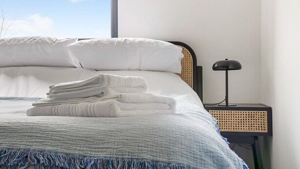 A neatly made bed with white linens and a blue fringe, a bedside lamp, and a view of the sky through a window.