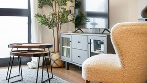 A modern living room with a grey cabinet, a fluffy chair, a small wooden table, and a large window with a view of trees.