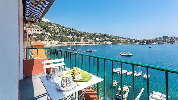 A balcony with a table set for breakfast, overlooking a marina with boats and a coastal town. - MAISON BLEUE AP4362 By Riviera Holiday Homes (Villefranche-sur-Mer)