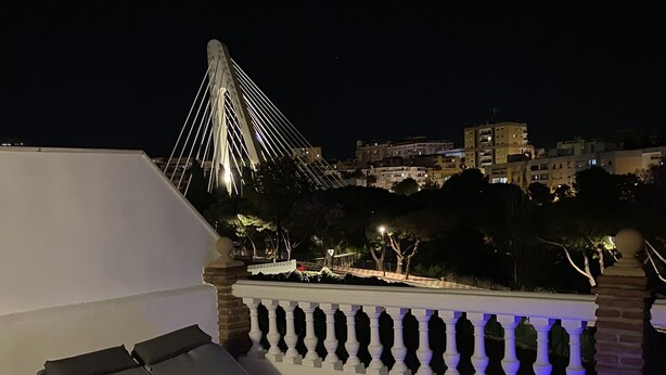 A rooftop terrace with a lounge chair and a red chair, a white balustrade, and a view of a bridge and buildings at night. - Casa Hemingway - Marbella Old Town (Marbella)