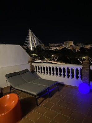Une terrasse sur le toit, avec un fauteuil de relaxation et une chaise rouge, une balustrade blanche, et une vue sur un pont et des bâtiments la nuit.