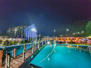 Vista nocturna de un hotel con una piscina grande, una terraza de madera y un edificio de fachada azul.