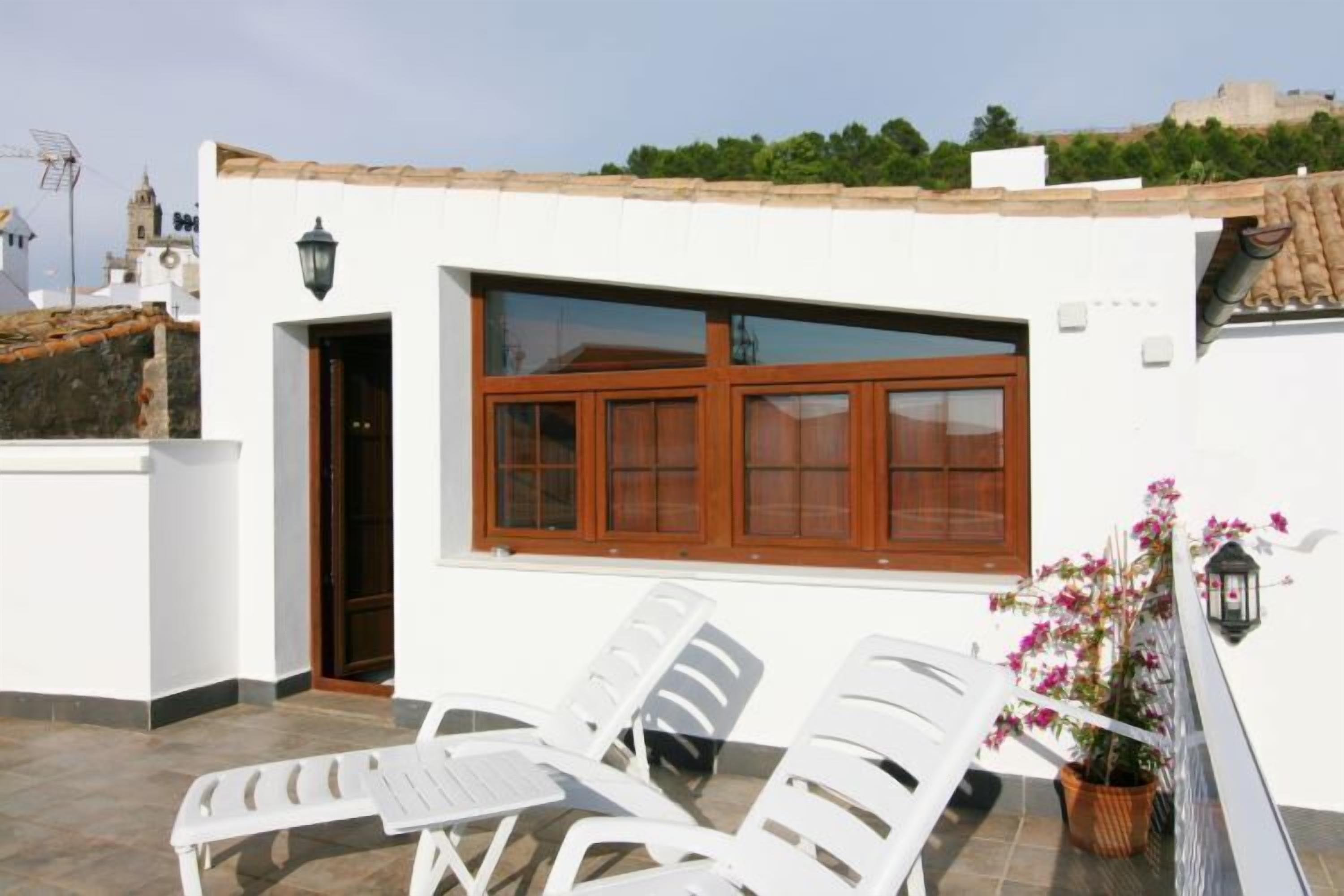 Un edificio blanco con techo de tejas, un ventanal con contraventanas de madera y un balcón con sillas de plástico blanco y una planta en maceta.