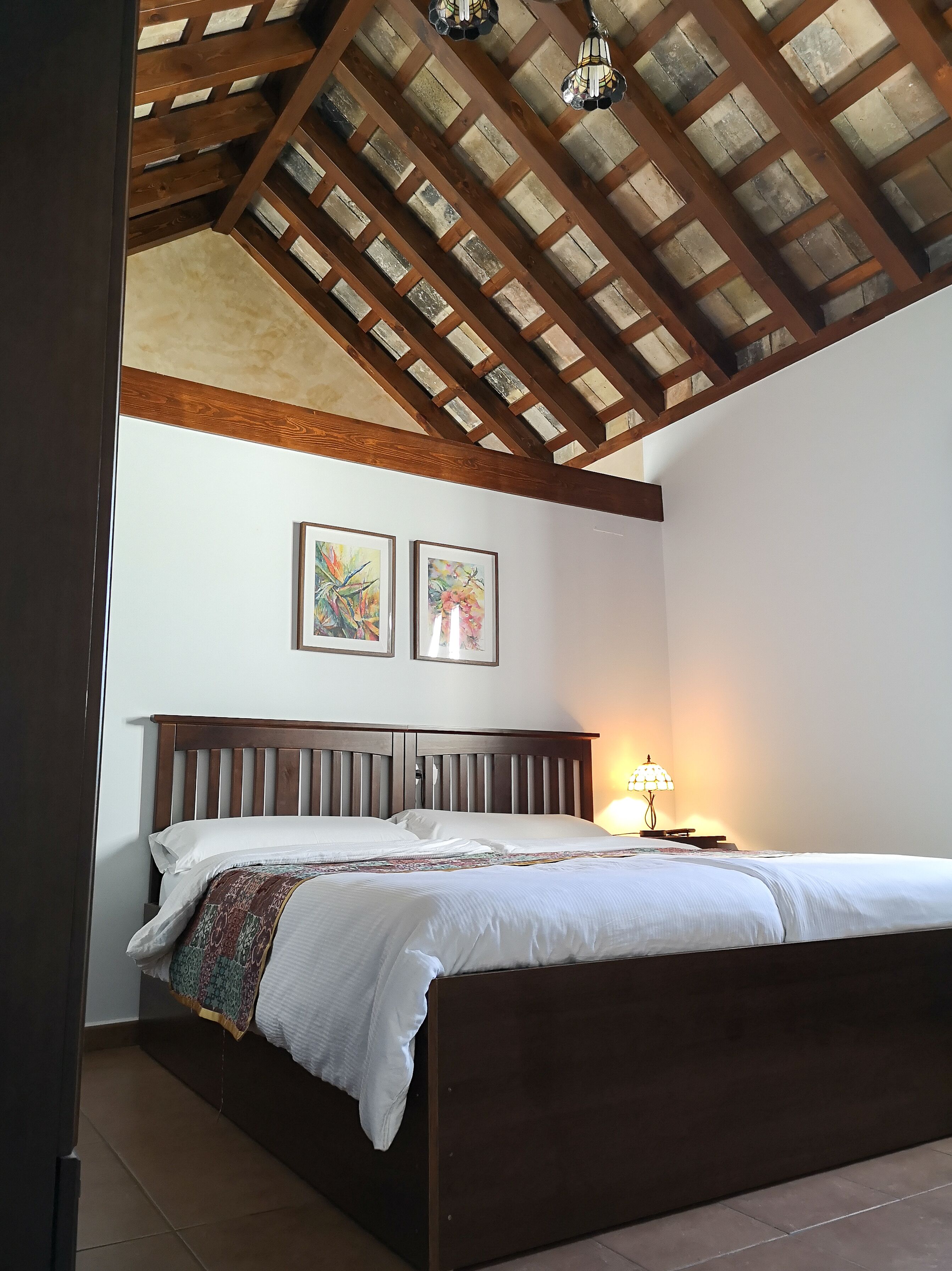 A bedroom with a wooden bed, two framed pictures on the wall, and a ceiling with exposed beams.