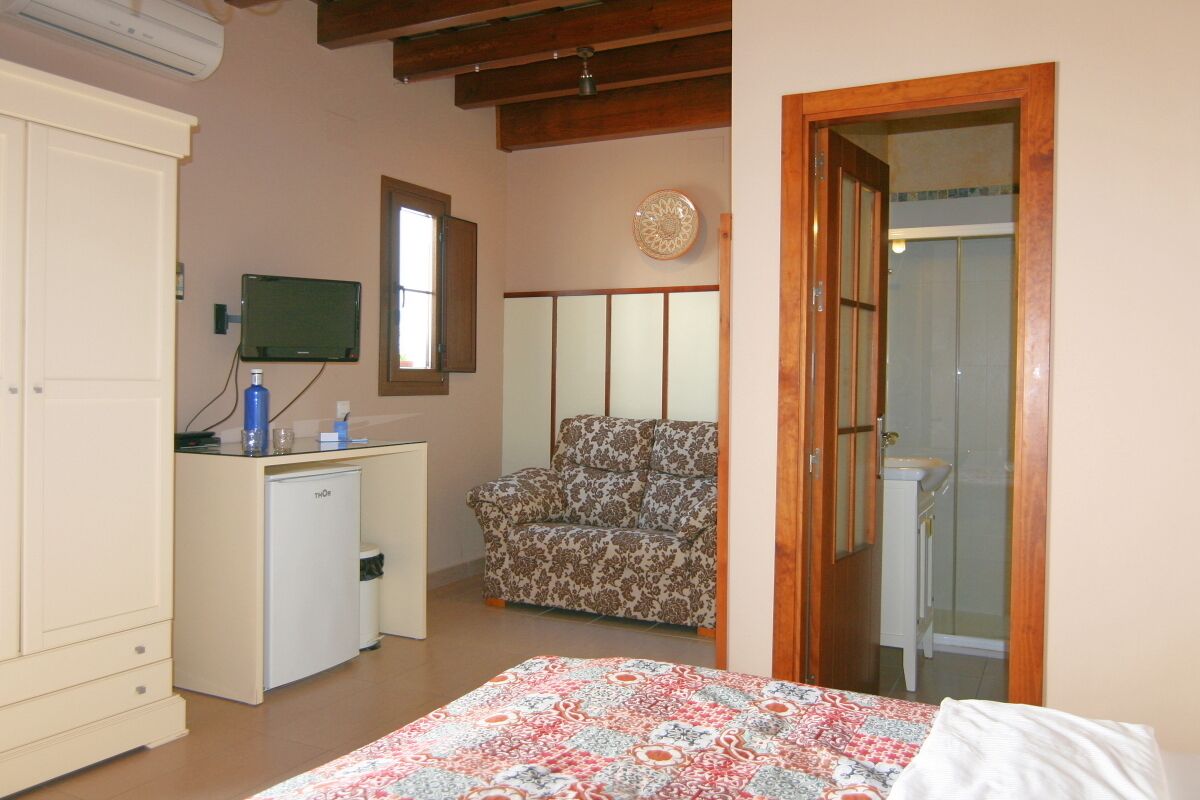 Un dormitorio con cama, escritorio con computadora, silla, armario y baño visible a través de una puerta abierta.