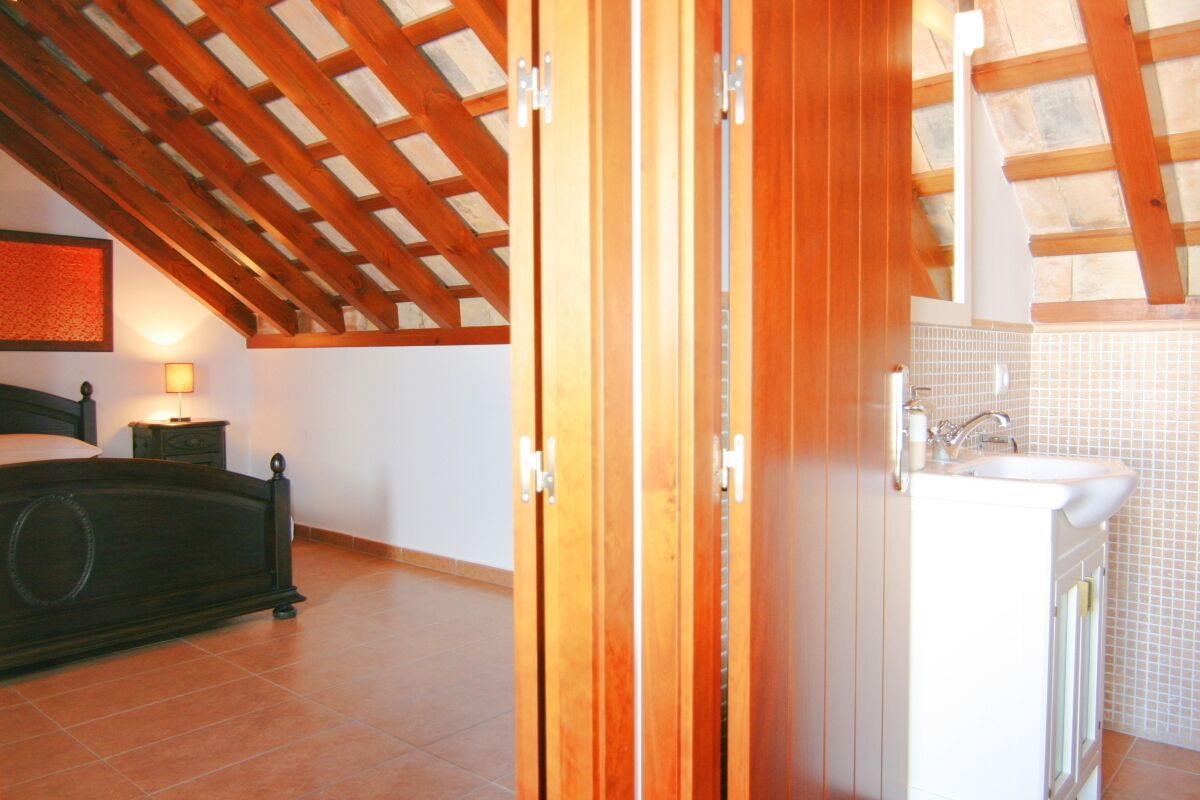 A bedroom with a wooden ceiling, a bed, a nightstand, and a bathroom with a sink and tiled walls.