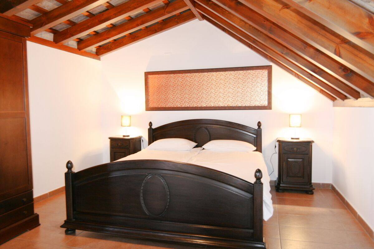 A bedroom with a wooden ceiling, a bed with a headboard, two bedside tables, and a wall-mounted light fixture.