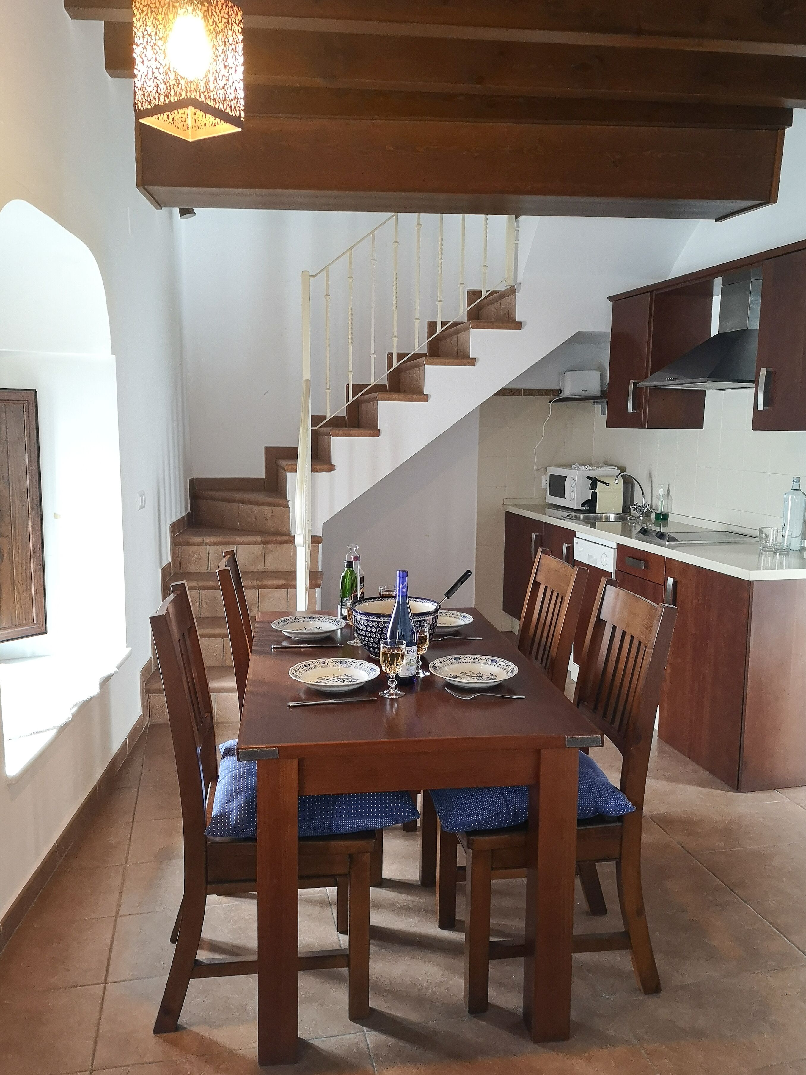 A dining area with a wooden table set for six, surrounded by chairs with blue cushions. A staircase leads to an upper level, and a kitchen area is visible to the right.