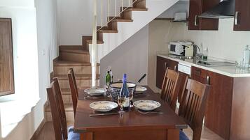 A dining area with a wooden table set for six, surrounded by chairs with blue cushions. A staircase leads to an upper level, and a kitchen area is visible to the right.