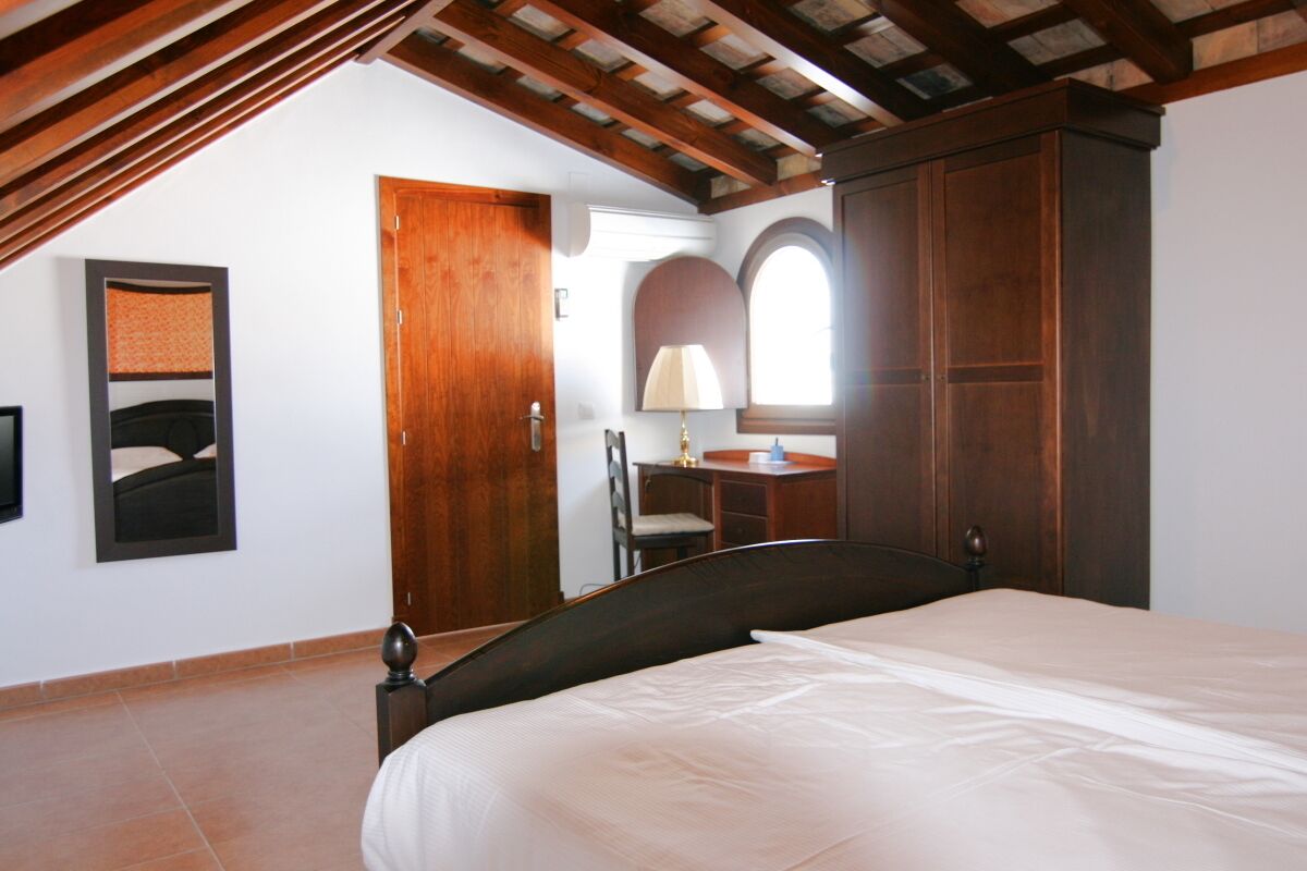 A bedroom with a large bed, wooden wardrobe, and a desk with a lamp.