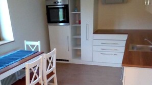 Kompakti keittiö, jossa on valkoiset kaapit, sisäänrakennettu uuni ja puinen ruokapöytä tuoleineen.