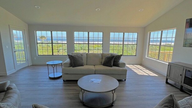Een ruime woonkamer met een groot raam met uitzicht op een strand, een ronde salontafel en een bank met kussens.