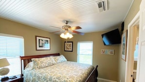 Un dormitorio con cama, mesita de noche, ventilador de techo y televisor montado en la pared.