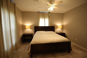 Une chambre à coucher avec un lit, deux tables de chevet, un ventilateur de plafond et des rideaux.