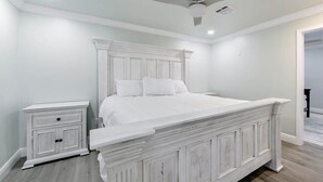 A bedroom with a white bed, a nightstand, a ceiling fan, and a door leading to another room.