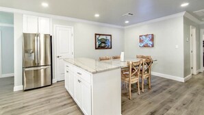 A modern kitchen with white cabinetry, a stainless steel refrigerator, a marble island, and wooden chairs.